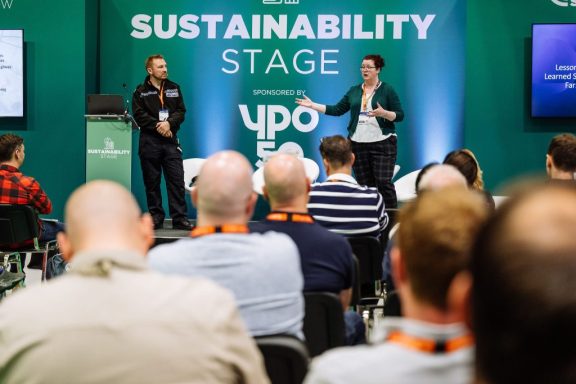 Two speakers presenting on stage at the Sustainability Stage, with an audience in view.